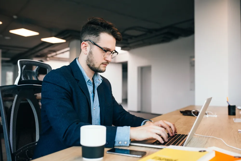 young dark haired man is working at the table in office. he wears blue shirt with black jacket. he is typing on laptop and looks busy